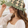Sage green crochet visor cap with ivory five-petal daisies and butter-yellow centers, worn by woman with brown wavy hair